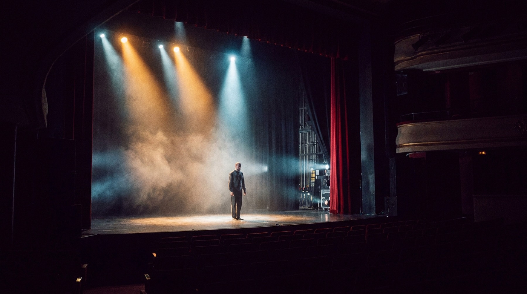 Un plateau de théâtre traversé par trois faisceaux de lumière, une silhouette d'artiste au centre